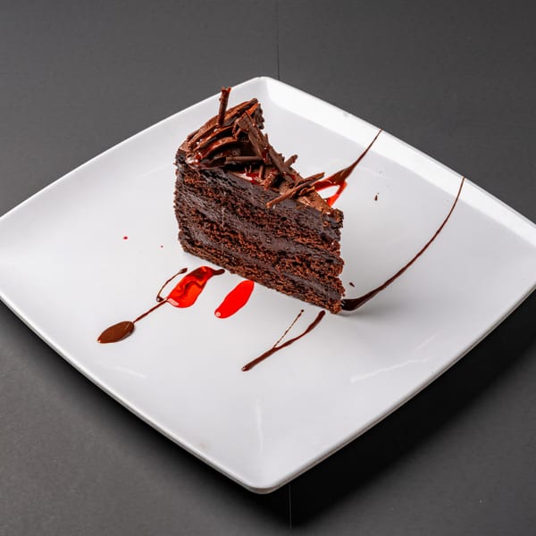 Slice of Bella's rich chocolate cake with chocolate shavings and raspberry drizzle on a white plate.