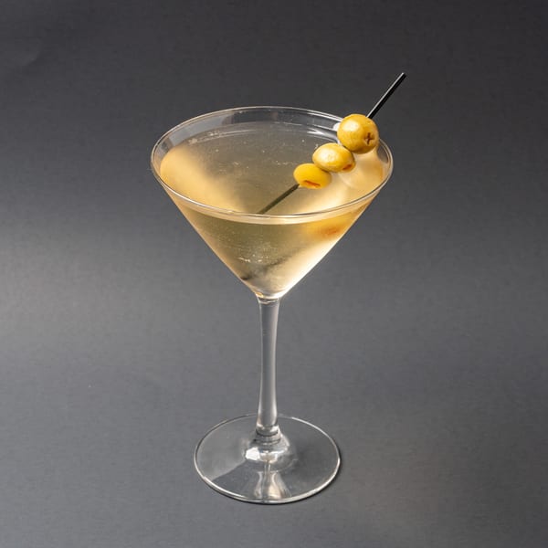 Martini cocktail with three green olives on a skewer, against a dark gray background.