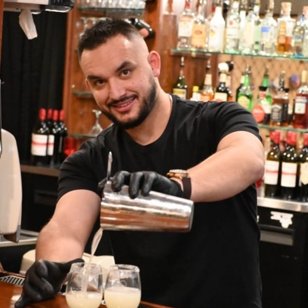 Smiling bartender in black gloves pouring a drink from a shaker into two glasses at Bella's Italian Restaurant.