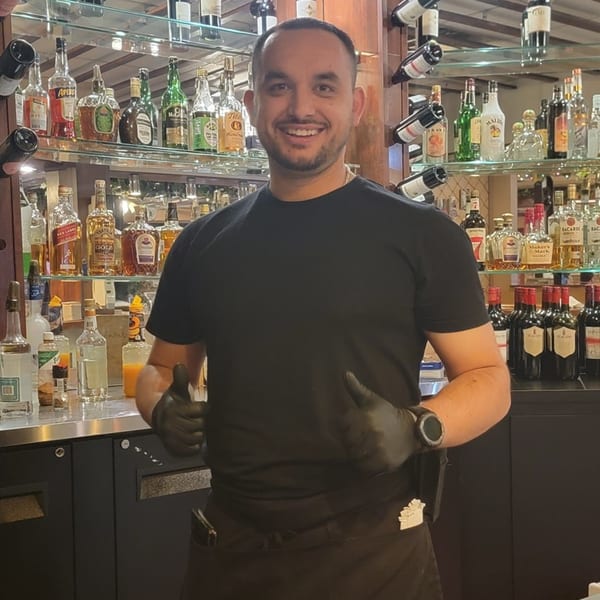 Smiling Bella's Italian Restaurant bartender in black shirt and gloves, giving thumbs up behind a bar.