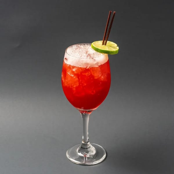 A vibrant red cocktail with foam, ice, a lime slice, and two brown straws in a wine glass.